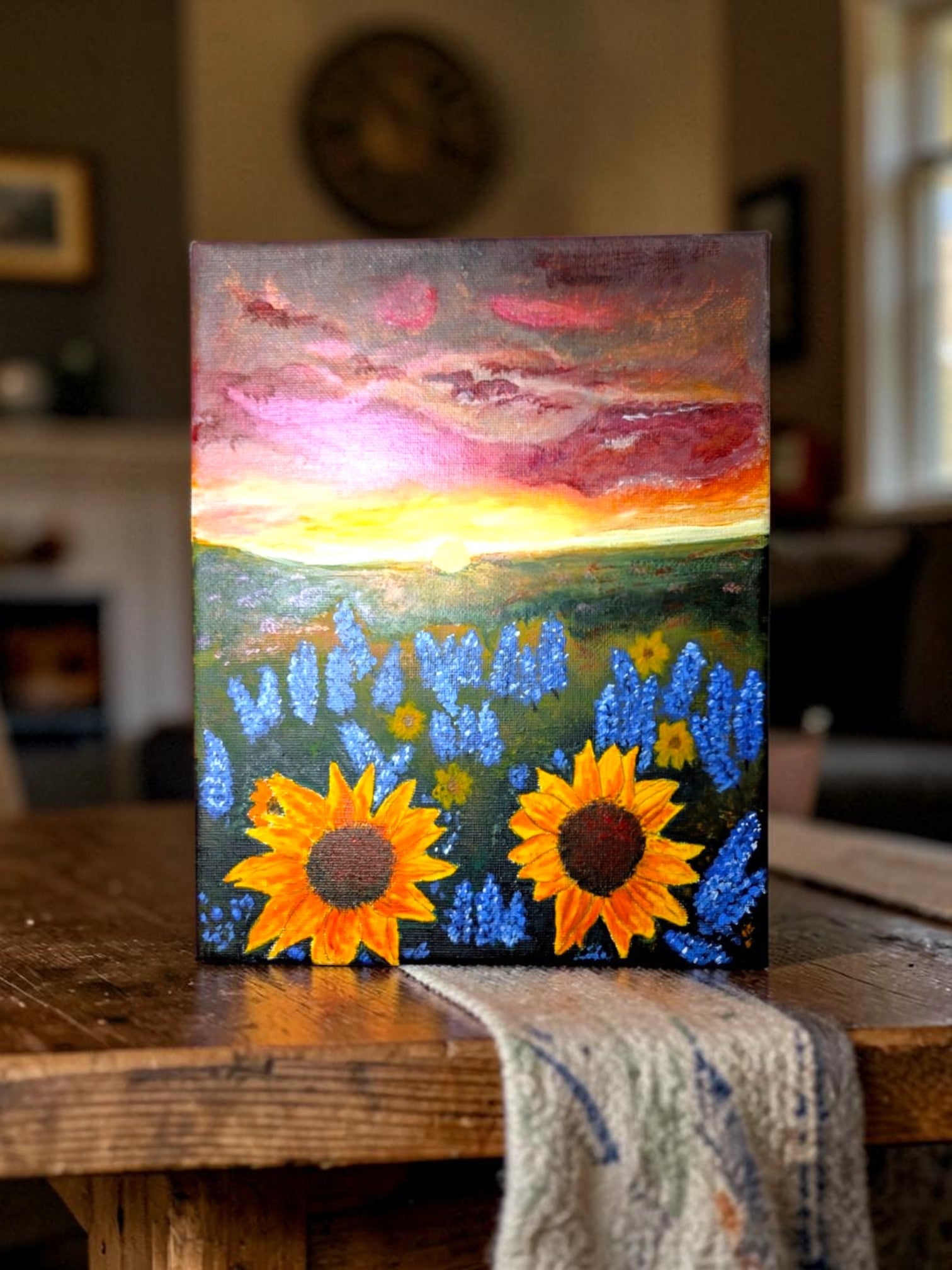 Hand-painted acrylic landscape of vibrant yellow sunflowers and purple bluebonnets in a field under a dramatic Texas sunset with shades of orange and pink.