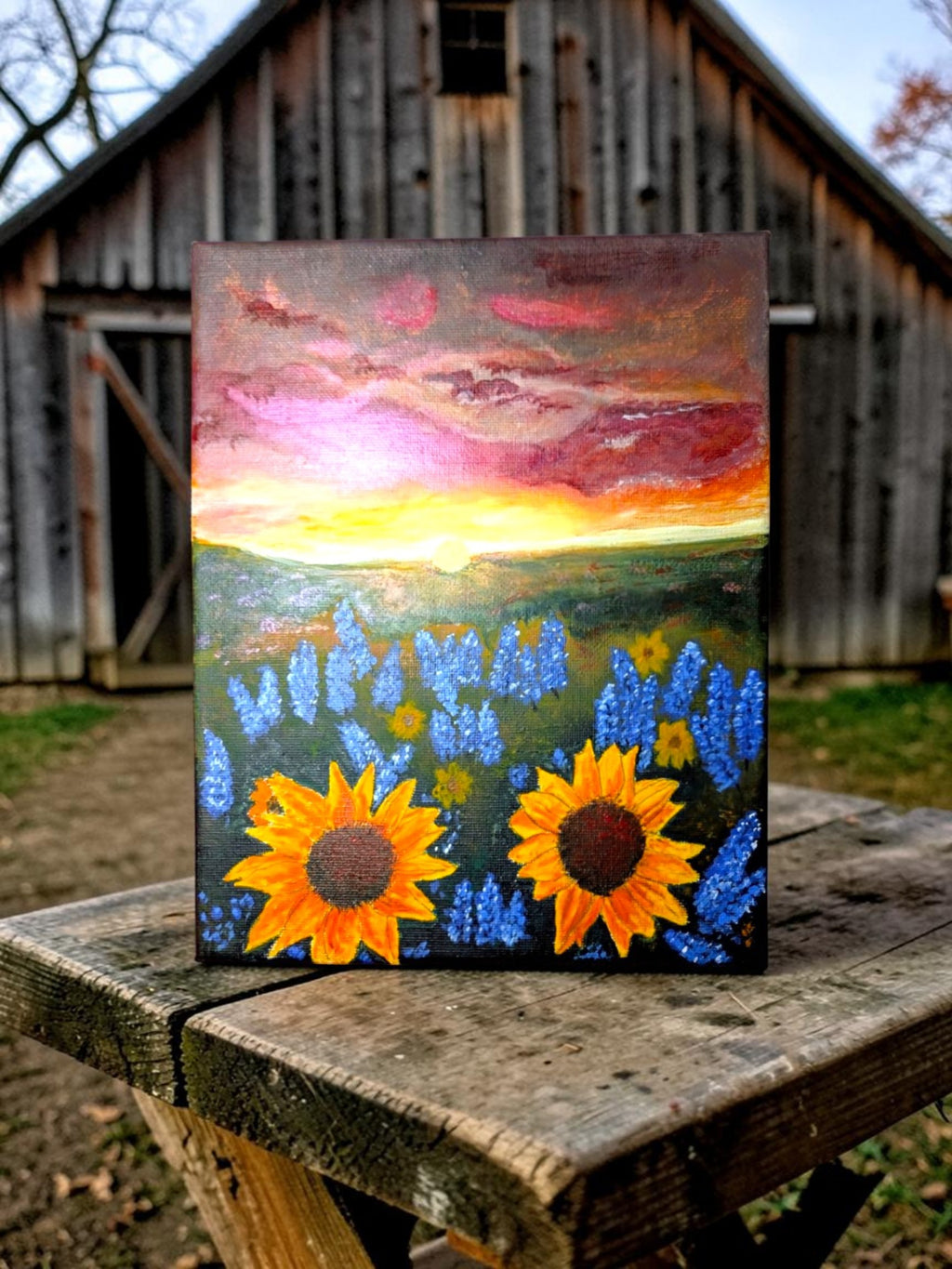 Hand-painted acrylic landscape of vibrant yellow sunflowers and purple bluebonnets in a field under a dramatic Texas sunset with shades of orange and pink.